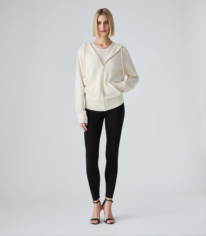A woman stands against a light gray studio background wearing a cream zip-front hoodie over a white top, black leggings, and black heeled sandals.