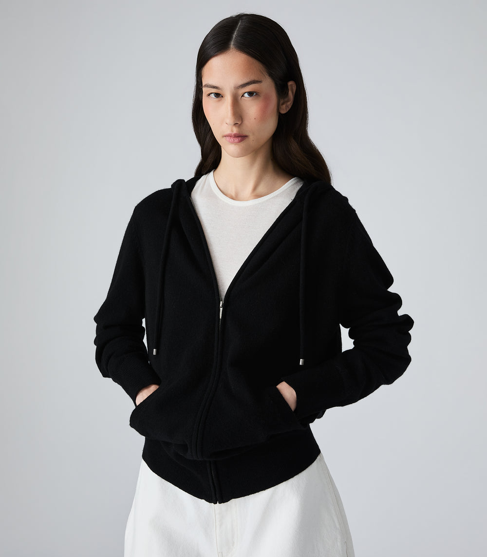 A woman modeling a black zip-up hoodie with drawstrings and front pockets, layered over a white crewneck top and white bottoms, against a plain light gray studio background.