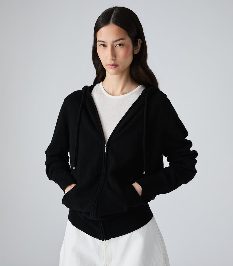 A woman modeling a black zip-up hoodie with drawstrings and front pockets, layered over a white crewneck top and white bottoms, against a plain light gray studio background.