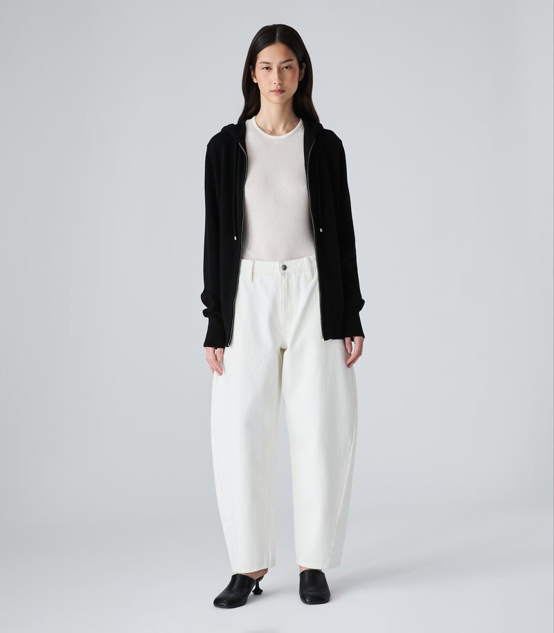 A woman modeling a black zip-up hoodie with drawstrings and front pockets, layered over a white crewneck top and white bottoms, against a plain light gray studio background.