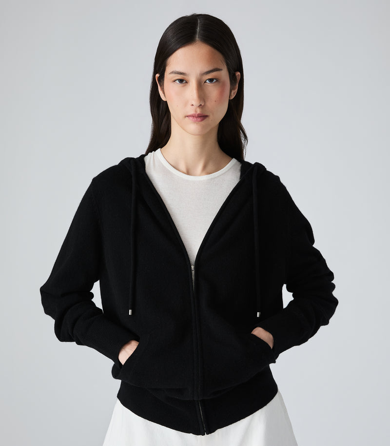 A woman modeling a black zip-up hoodie with drawstrings and front pockets, layered over a white crewneck top and white bottoms, against a plain light gray studio background.