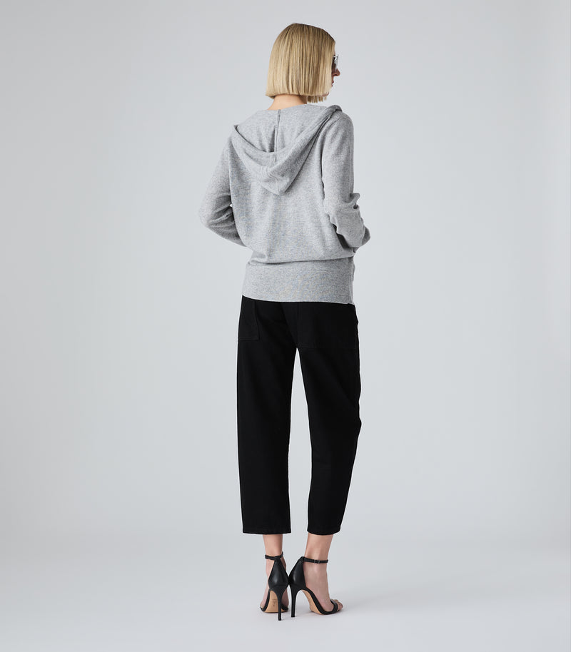 A woman is shown from behind wearing a light gray heathered hooded sweatshirt and black cropped trousers, paired with black high-heeled sandals, against a light gray studio background.