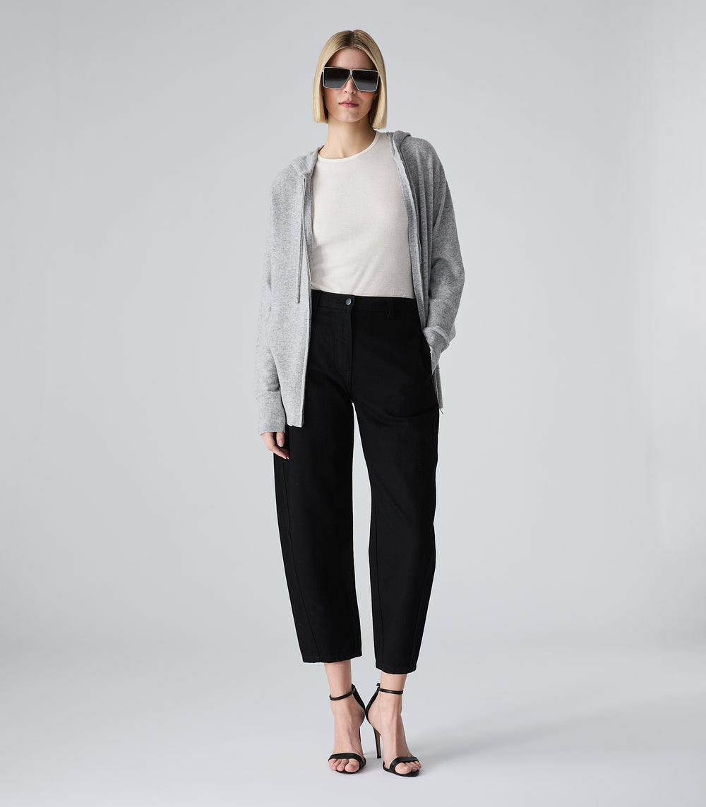 A model stands against a light gray studio background wearing a light gray open-front cardigan over a cream top, black cropped trousers, and black strappy heels.