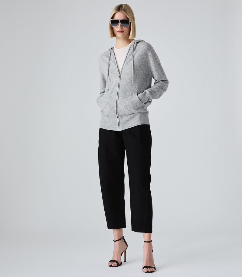 A model wearing a light gray zip-up hoodie, black cropped trousers, and black high-heeled sandals, standing against a light gray studio background.