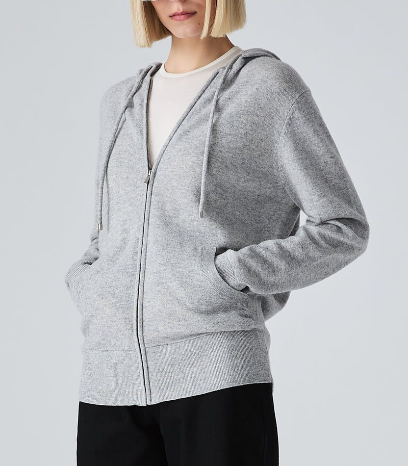 A woman wearing a light gray heather zip-up hoodie over a white crewneck shirt and black pants, shown from the chin down against a light gray studio background.