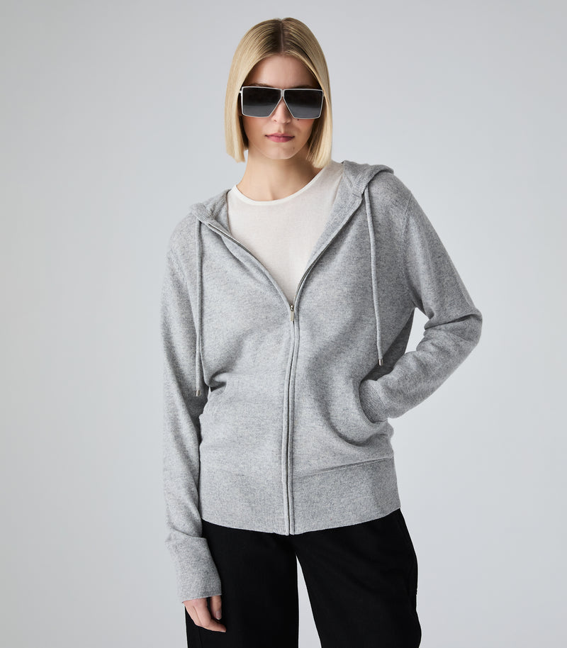 A woman with a short blonde bob and sunglasses wearing a light gray zip-up hoodie over a white crewneck top and black pants, standing against a light gray studio background.