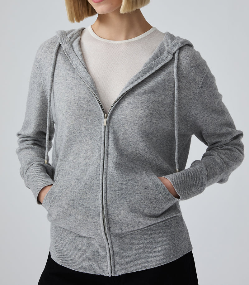 A woman wearing a heather gray zip-up hoodie over a cream crewneck top and black bottoms, shown from the chin down against a light gray studio background.
