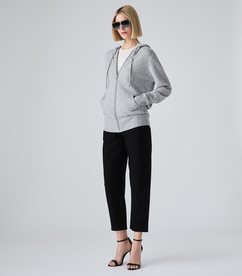 A woman modeling a light gray zip-up hoodie with black cropped pants and black strappy heels, standing against a light gray studio background.