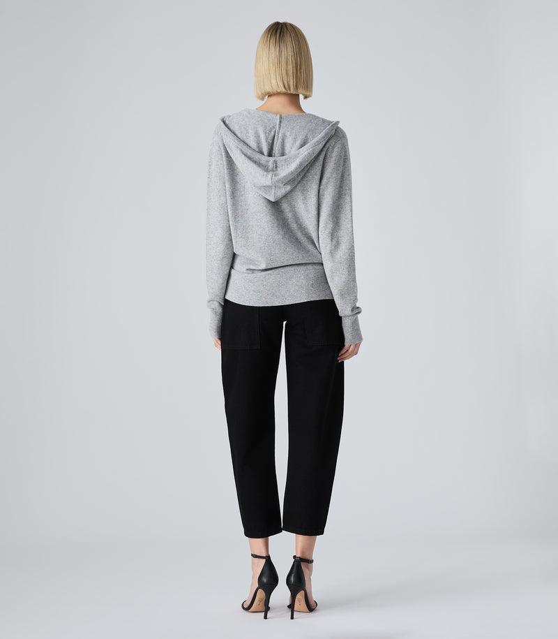 A woman viewed from the back wearing a light gray heathered hooded sweatshirt, black cropped pants, and black strappy heels against a light gray studio background.