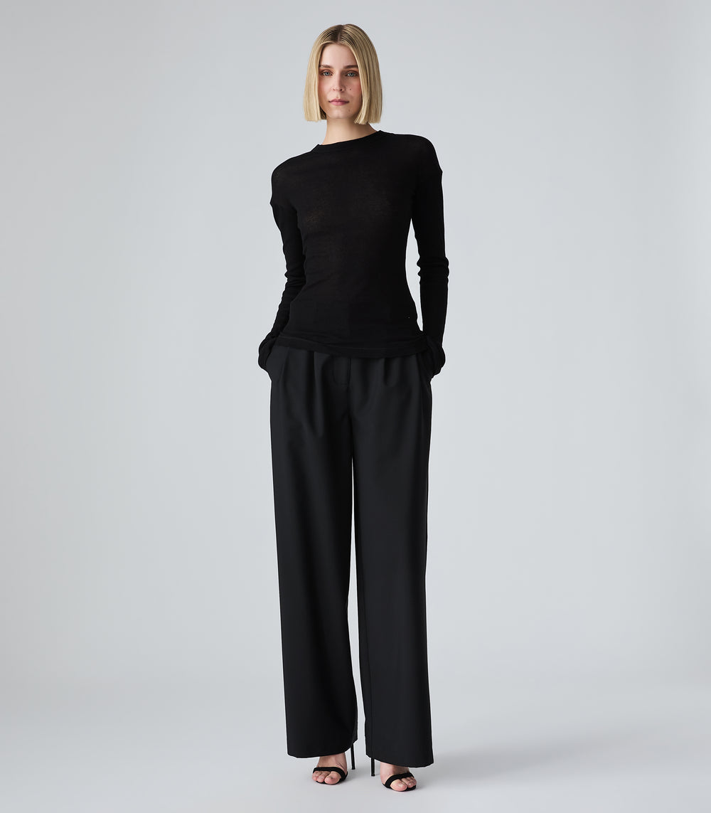Full-length view of a model wearing high-rise, wide-leg black trousers featuring a relaxed, fluid silhouette and delicate front pleat details at the waist. The trousers are styled with both hands in the pockets, paired with a fitted black crew-neck long-sleeved top and black stiletto heels against a light grey background.