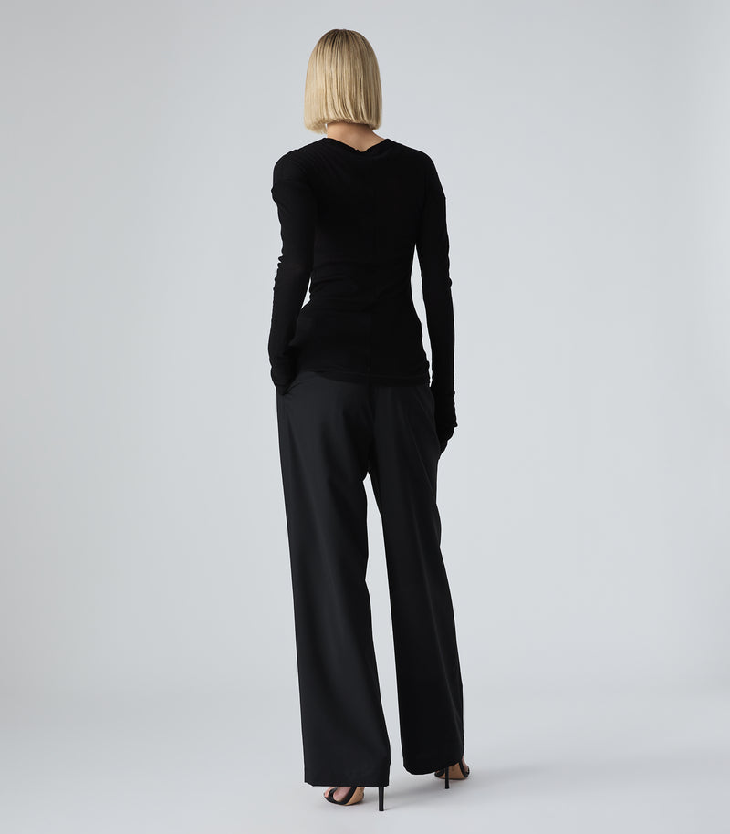 Full-length rear view of a model wearing high-rise, wide-leg black trousers featuring a relaxed, fluid silhouette with clean draping and horizontal slip back pockets. The trousers are styled with a fitted black long-sleeved fine-knit top and black stiletto heels against a light grey background.