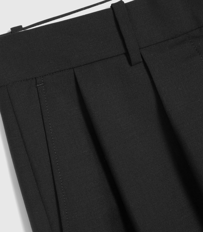 Macro close-up view of the upper waistband on a pair of black tailored trousers, highlighting the fine wool-blend fabric texture. The shot focuses on a sharp vertical front pleat, a structured fabric belt loop, and a subtly stitched side slip pocket opening.