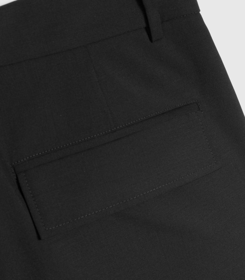 Macro close-up view of the back pocket on a pair of black tailored trousers, highlighting the fine wool-blend fabric texture. The shot focuses on a structured welt pocket featuring a sharp rectangular flap with neat tonal topstitching, positioned just below a fabric belt loop on the waistband.
