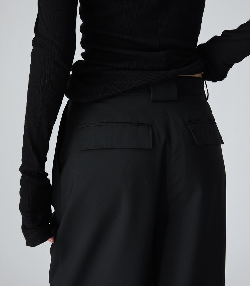 Close-up rear view of a model wearing high-rise, wide-leg black trousers featuring a relaxed silhouette, horizontal welt pockets with sharp rectangular flaps at the seat, and a wide central belt loop. The trousers are styled with a fitted black long-sleeved ribbed knit top, which is slightly pulled up to showcase the tailored waistband details against a light grey background.