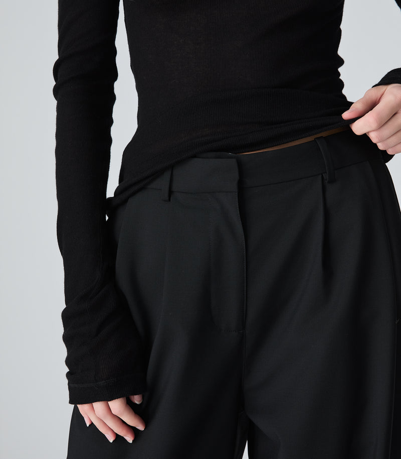 Close-up view of a model wearing high-rise, wide-leg black trousers featuring a relaxed silhouette, sharp front pleat detailing, structured belt loops, and an asymmetrical overlap waistband closure design. The trousers are styled with a fitted black long-sleeved fine-knit top, lifted slightly by a hand at the hip to reveal the tailored front waist construction against a light grey background.