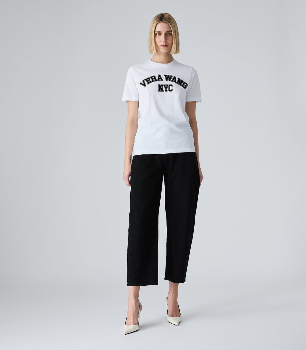 Full-length view of a model wearing high-rise, ankle-length cropped black jeans featuring a structured balloon silhouette and curved leg seams. The jeans are styled with a white varsity graphic t-shirt and white pointed-toe slingback heels against a light grey background.