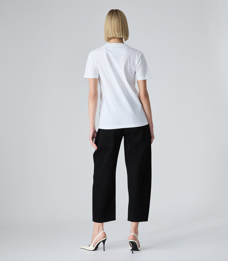 Full-length back view of a model wearing high-rise, ankle-length cropped black jeans featuring a structured balloon silhouette and curved leg seams. The jeans are styled with a white t-shirt and white pointed-toe slingback heels against a light grey background.