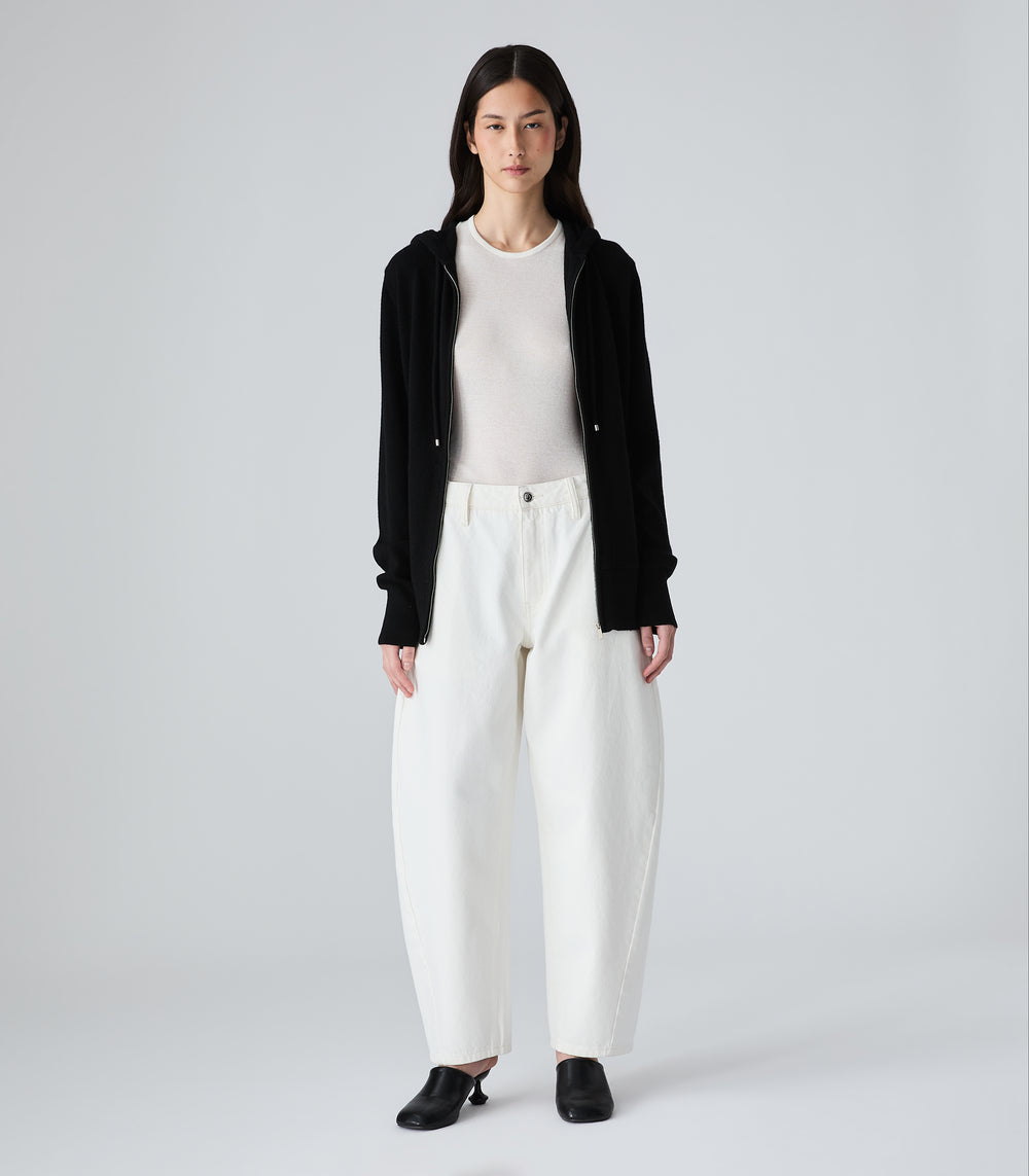 Full-length view of a model wearing high-waisted, wide-leg ecru jeans with a relaxed balloon silhouette, styled with a white top, open black zip-up hoodie, and black pointed-toe mule heels.