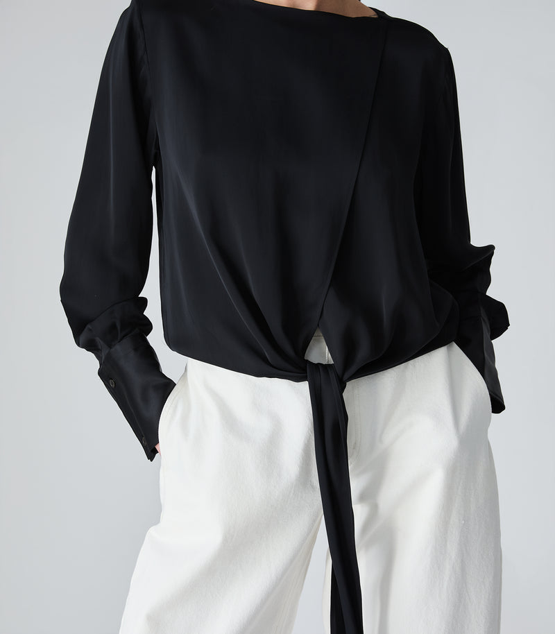 A person wearing a black long-sleeve wrap-style blouse with a side tie and white wide-leg pants, photographed against a light gray studio background.
