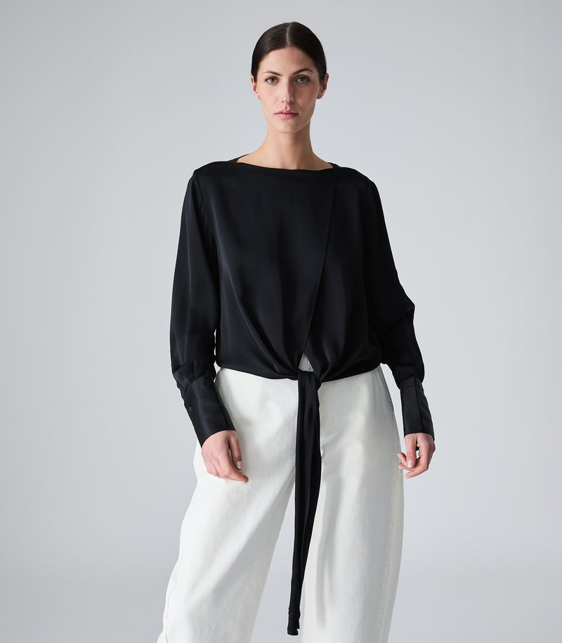A woman modeling a black long-sleeve wrap-style blouse with a tie detail at the waist, paired with white wide-leg pants, against a light gray studio background.