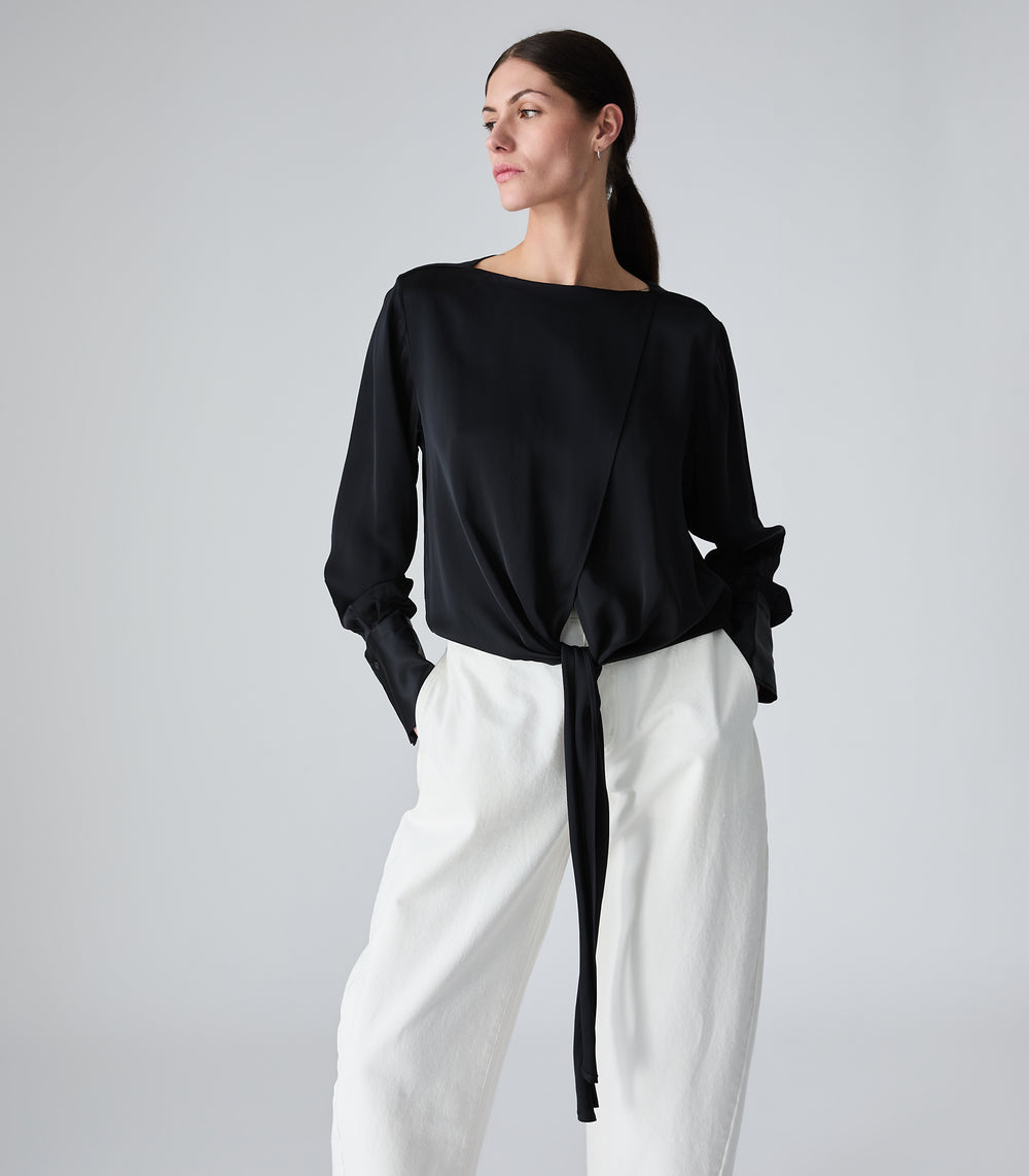 A woman modeling a black long-sleeve wrap-style top with a side tie detail, paired with white wide-leg pants, against a light gray studio background.