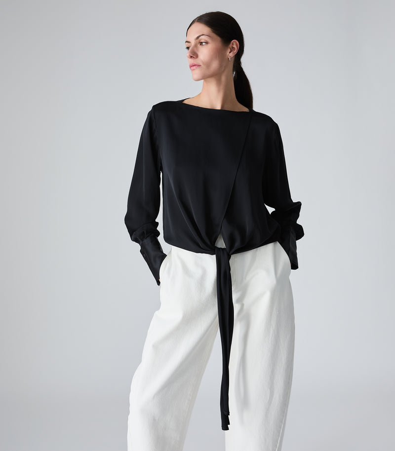 A woman modeling a black long-sleeve wrap-style top with a side tie detail, paired with white wide-leg pants, against a light gray studio background.