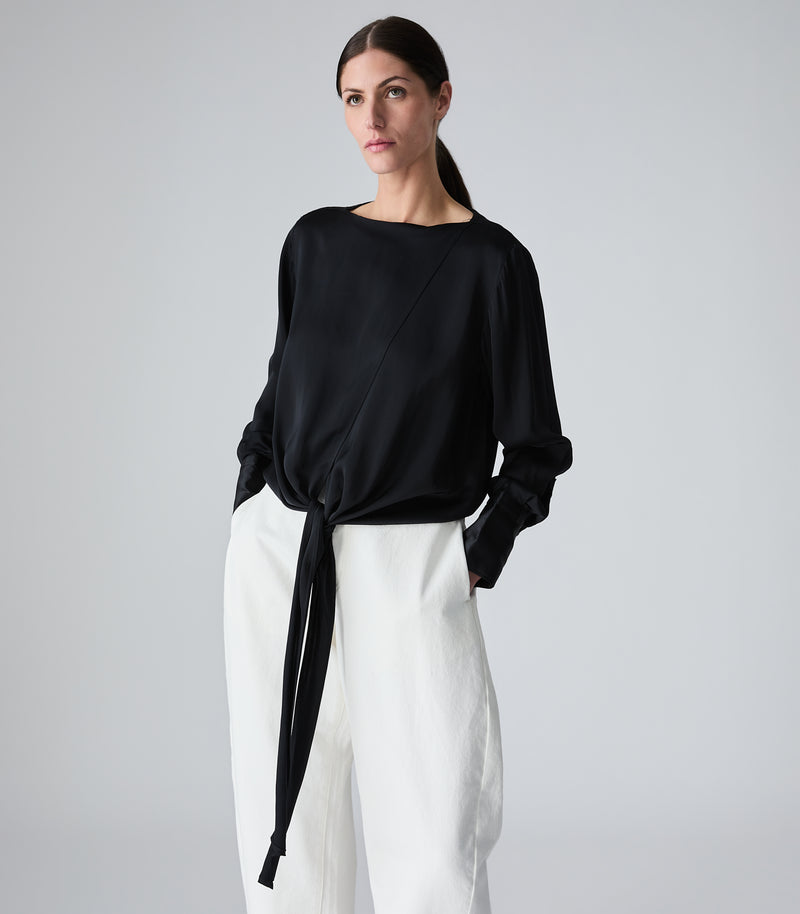 A woman modeling a black long-sleeve top with diagonal seam details and a front tie knot, paired with white wide-leg trousers, against a light gray studio background.