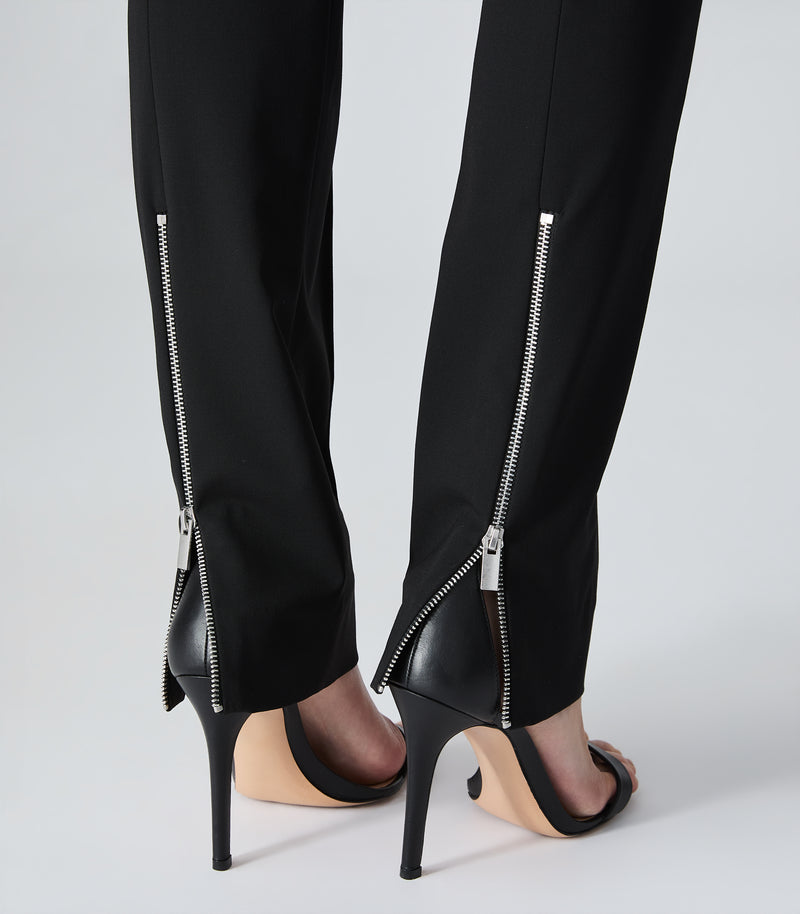 Black trousers with prominent silver zipper details running down the outer side of the lower legs, styled with black and nude high-heeled sandals against a light gray studio background.
