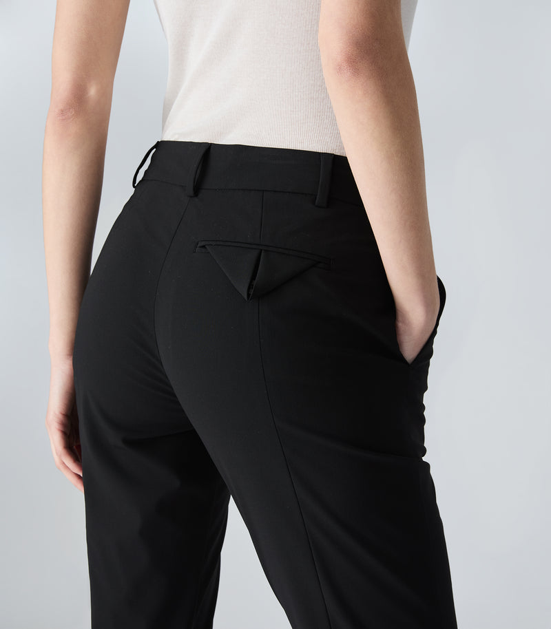Rear view of a person wearing black tailored trousers with a structured seat, belt loops, and a small triangular back pocket detail, paired with a light beige top against a light gray studio background.
