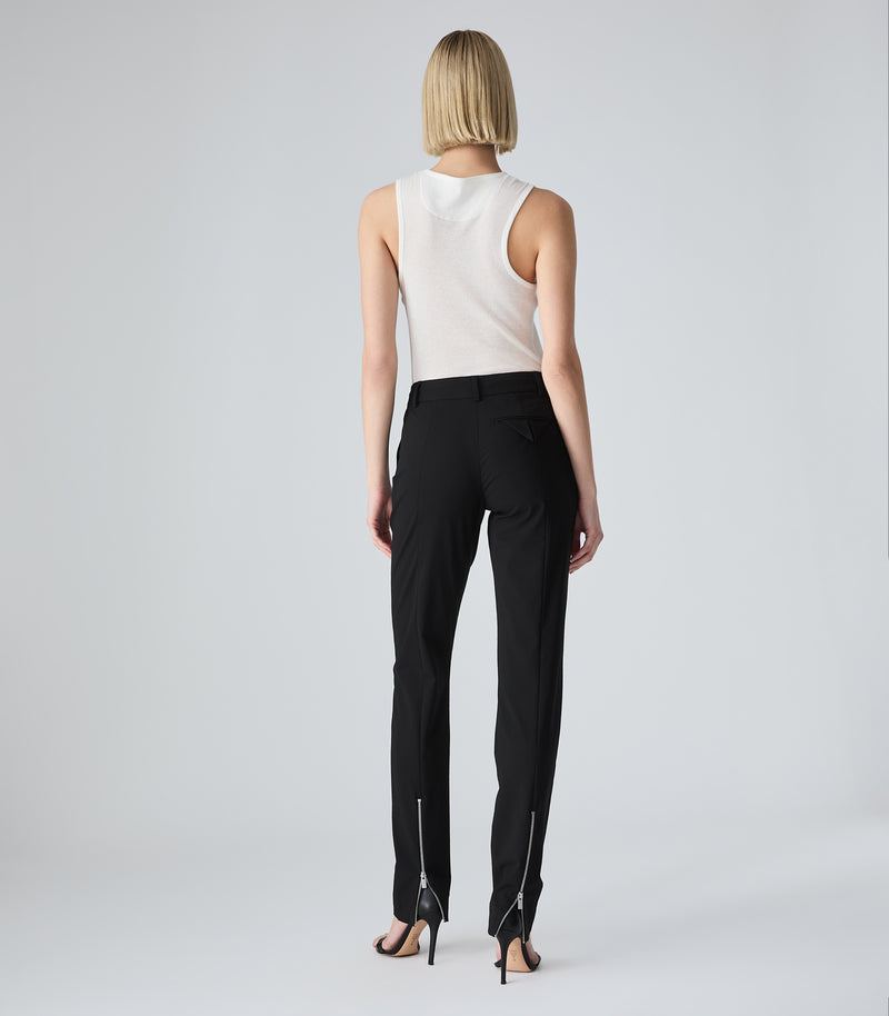 A model shown from the back wearing black trousers with silver zipper details along the outer sides, paired with a white sleeveless top and black high-heeled sandals, against a light gray studio background.