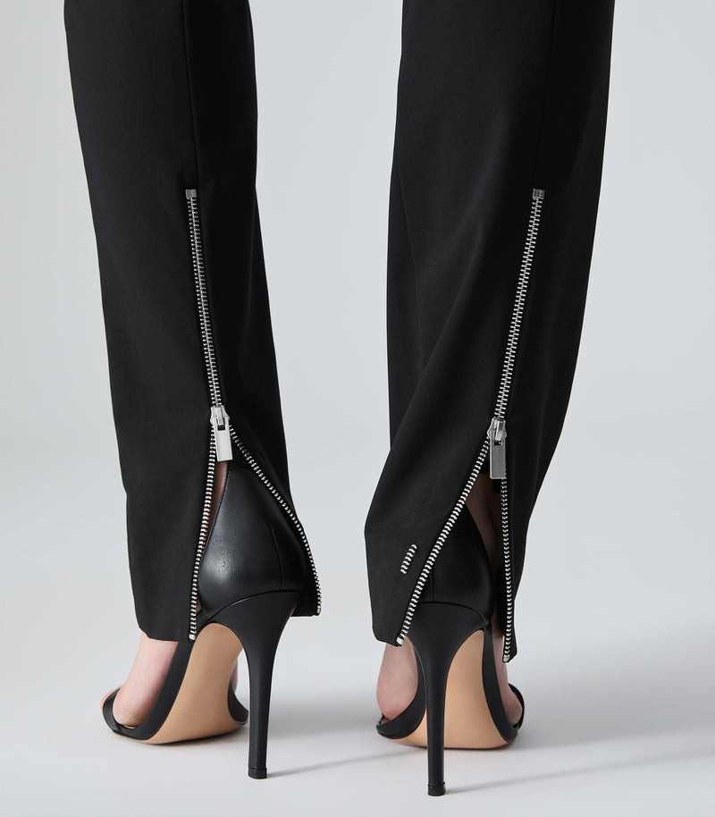 Close-up back view of a person wearing black trousers with silver side zippers and black high-heeled sandals on a light gray studio background.