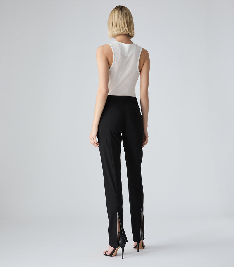 A model viewed from the back wearing a white sleeveless top and black high-waisted trousers with silver side zippers, paired with black high-heeled sandals, against a light gray studio background.
