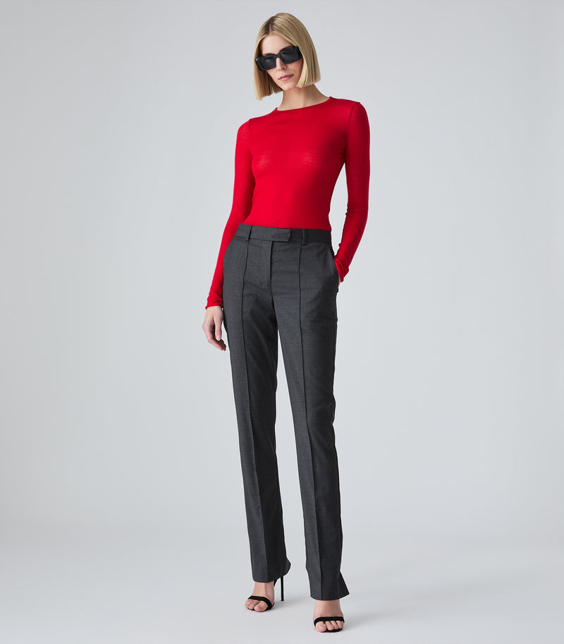 A woman stands in profile wearing a bright red long-sleeve knit top, dark charcoal gray trousers with side zippers, and black high-heeled sandals against a light gray studio background.