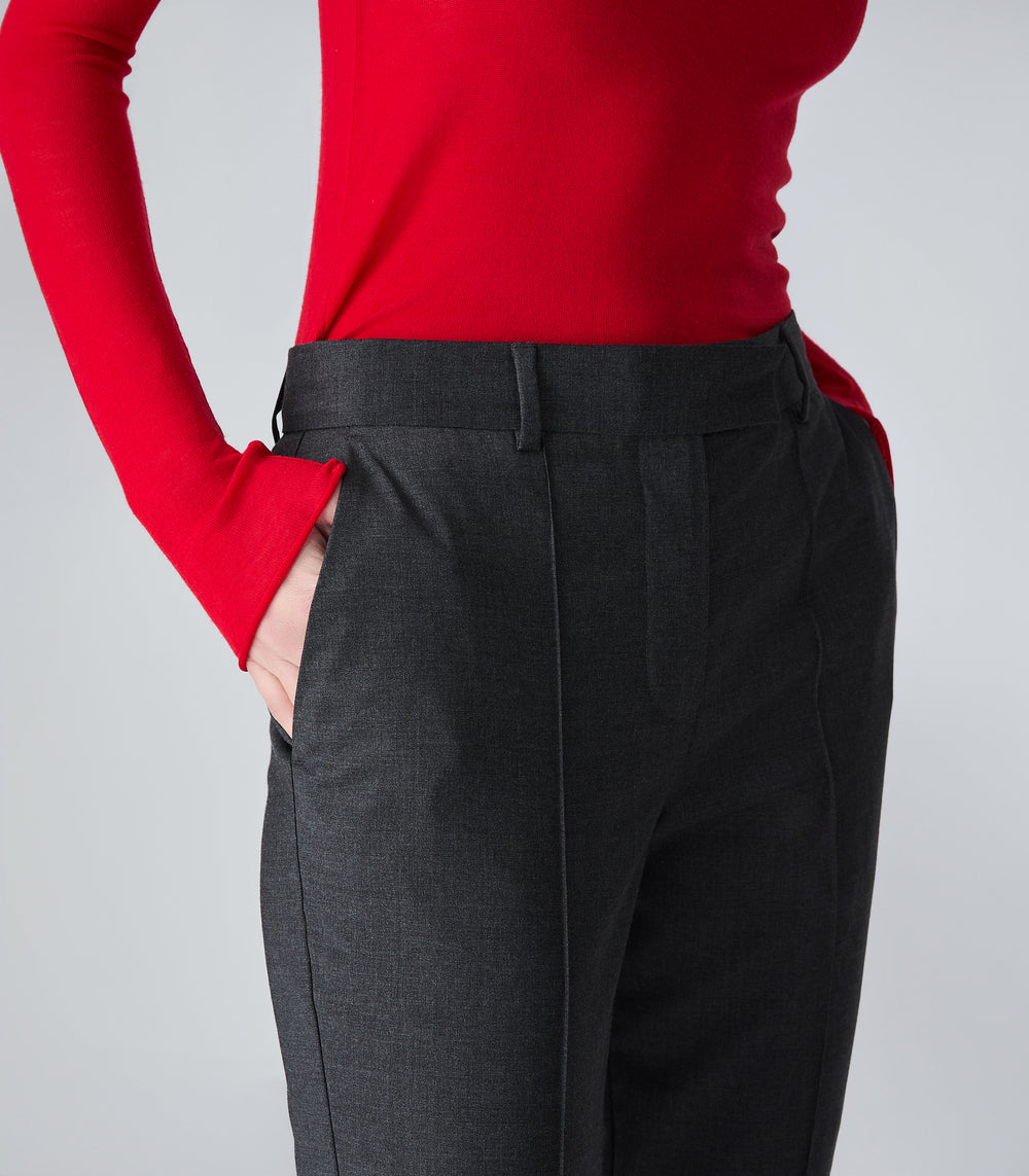 A close-up of a model wearing dark charcoal gray pleated trousers and a bright red long-sleeve top, with one hand in the trouser pocket, against a light gray studio background.