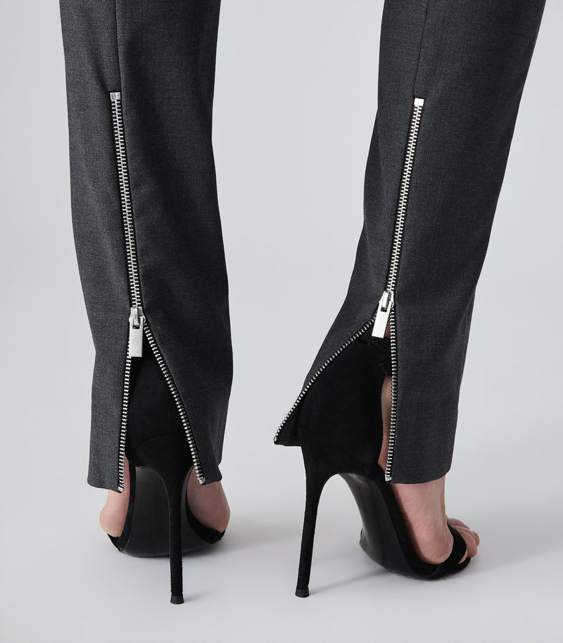 Close-up of a person's legs wearing dark gray trousers with silver zipper details at the ankles, paired with black high-heeled sandals against a light gray studio background.