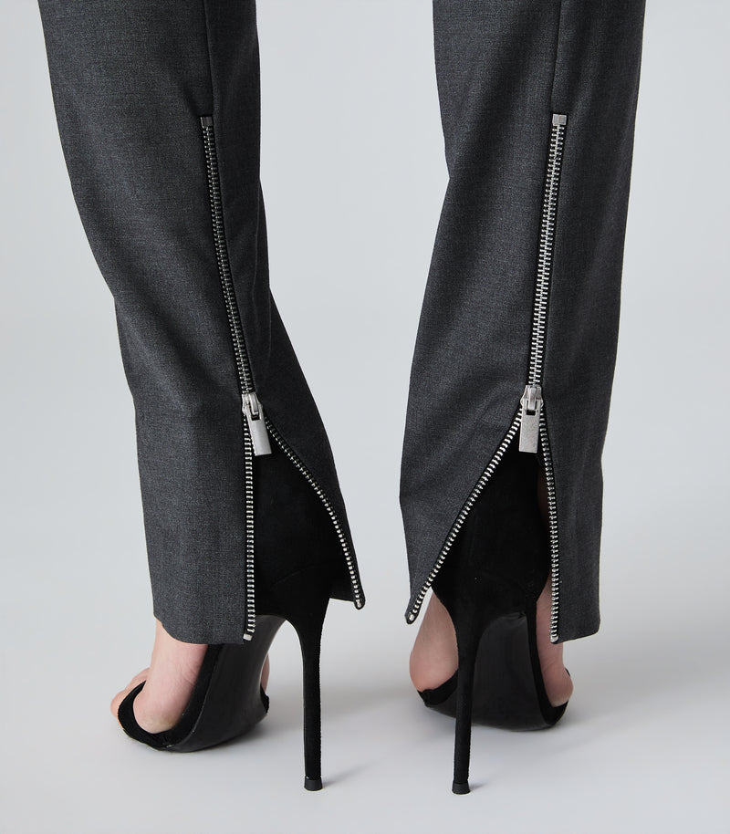 Close-up of a person's legs from the back, wearing dark gray wide-leg trousers with silver side zippers and black high-heeled sandals against a light gray studio background.
