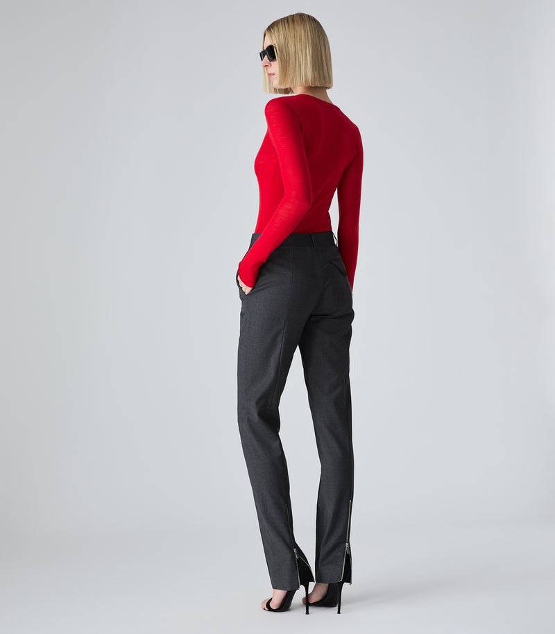 A woman stands in profile wearing a bright red long-sleeve knit top, dark charcoal gray trousers with side zippers, and black high-heeled sandals against a light gray studio background.