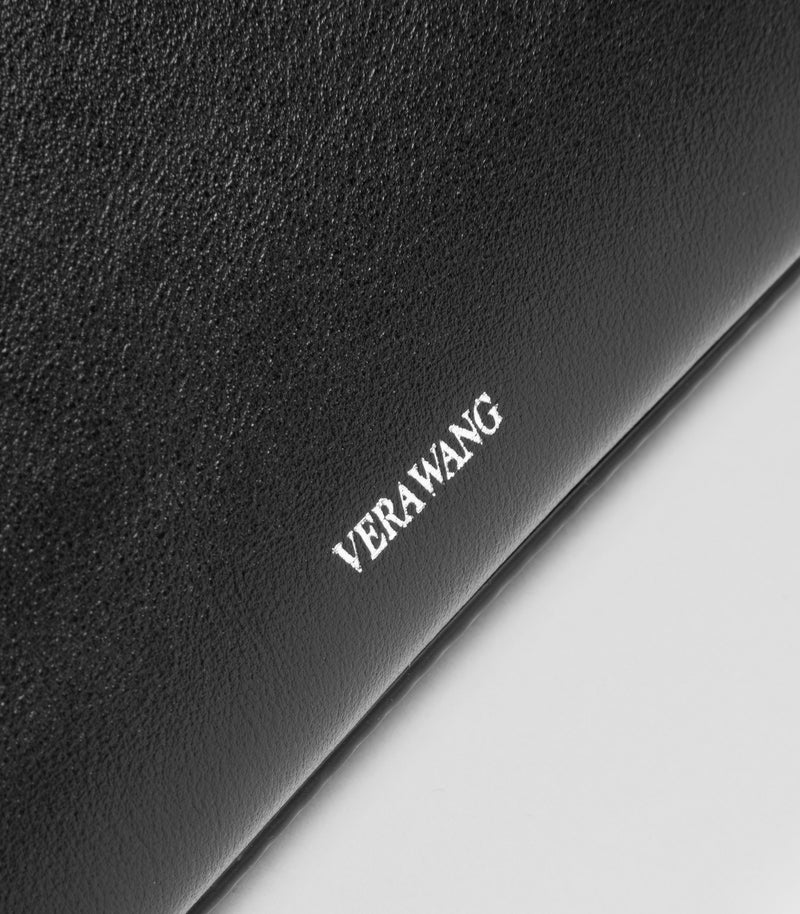 Close-up of a black textured leather handbag with the brand name 'VERA WANG' debossed in silver on the corner, set against a light gray background.