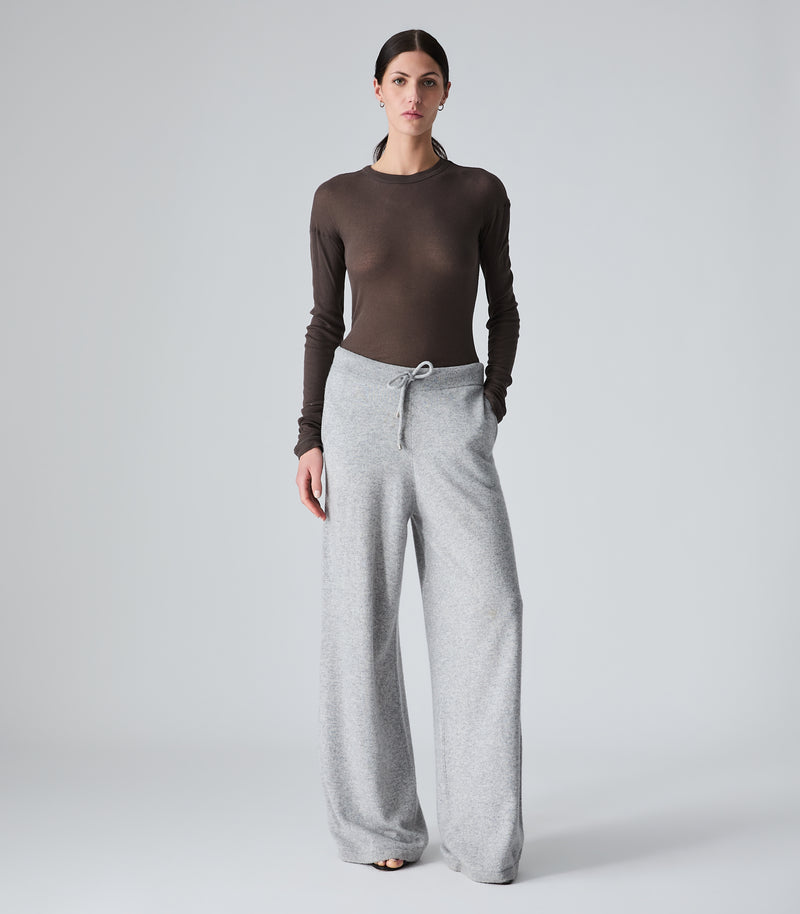 A woman modeling a dark brown long-sleeve mock neck top paired with light gray wide-leg sweatpants, standing against a neutral light gray studio background.