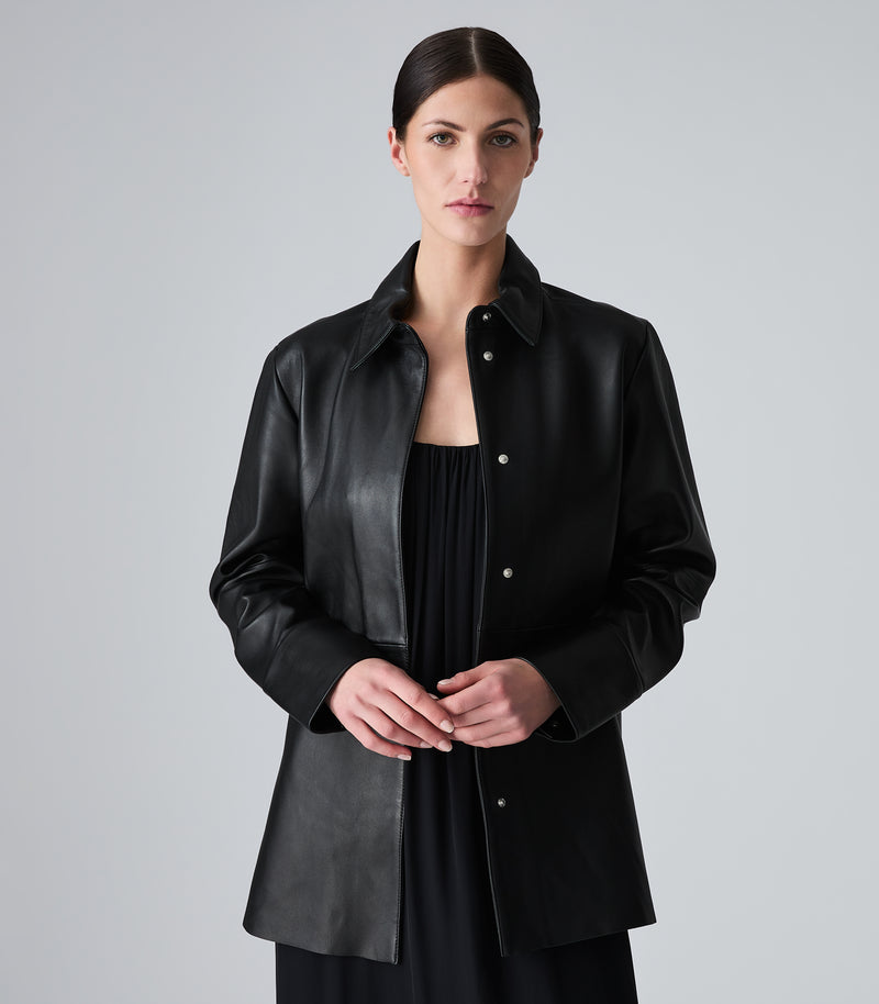 A woman wearing a black leather button-up shirt jacket over a black top, photographed against a neutral light gray studio background.