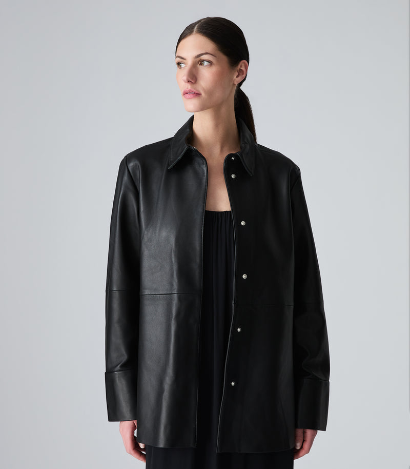 A woman wearing a black leather button-up shirt jacket over a black top, photographed against a neutral light gray studio background.