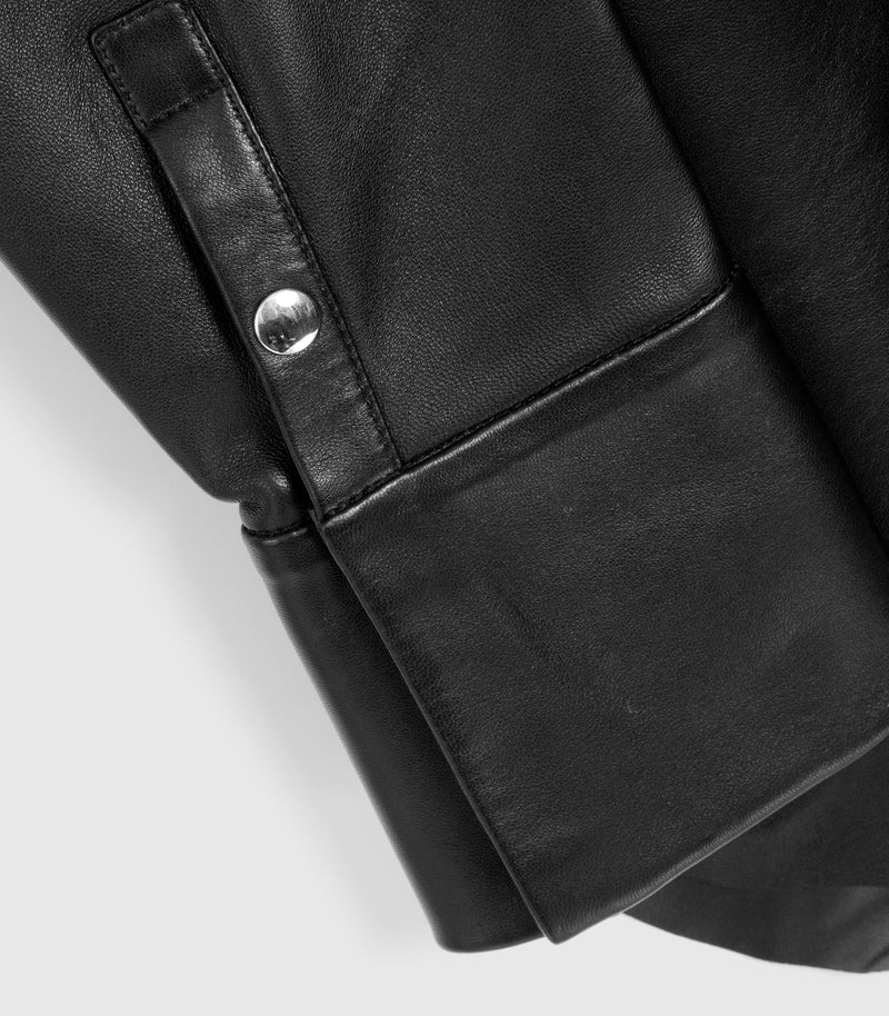 Close-up detail of a black leather jacket showing a sleeve cuff with a silver snap button closure, on a light gray background.