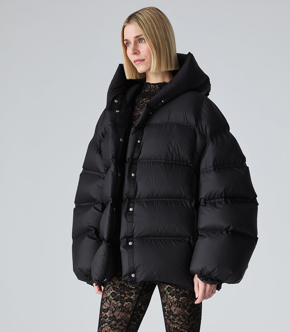 A woman modeling an oversized black puffer jacket with a high collar and snap-button front, paired with black lace leggings, against a light gray studio background.