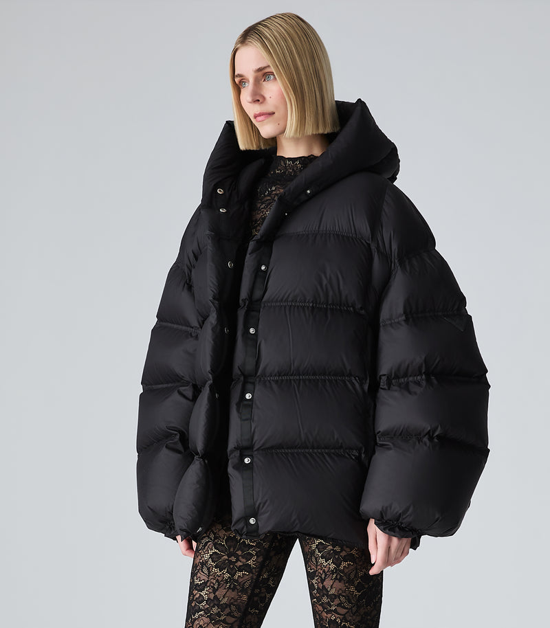 A woman modeling an oversized black puffer jacket with a high collar and snap-button front, paired with black lace leggings, against a light gray studio background.