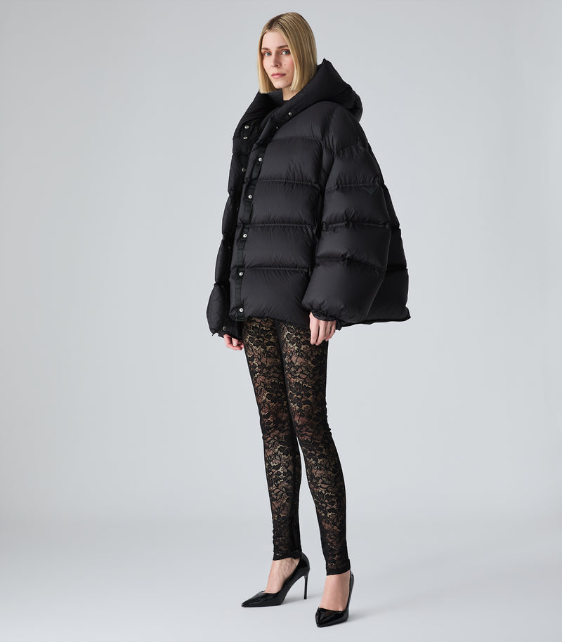 A woman modeling a black puffer jacket with a large black hood, paired with black lace leggings and black pointed-toe heels, against a light gray studio background.