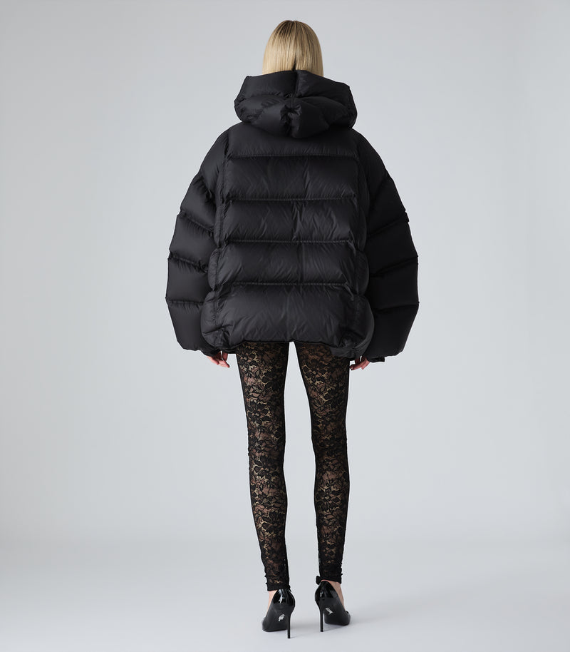 A model viewed from the back wearing a black oversized puffer jacket, black lace leggings, and black high heels against a light gray studio background.
