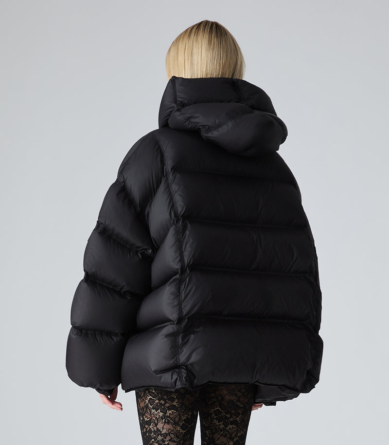 A person shown from the back wearing an oversized black puffer jacket with a high padded collar and horizontal quilting, against a light gray studio background.
