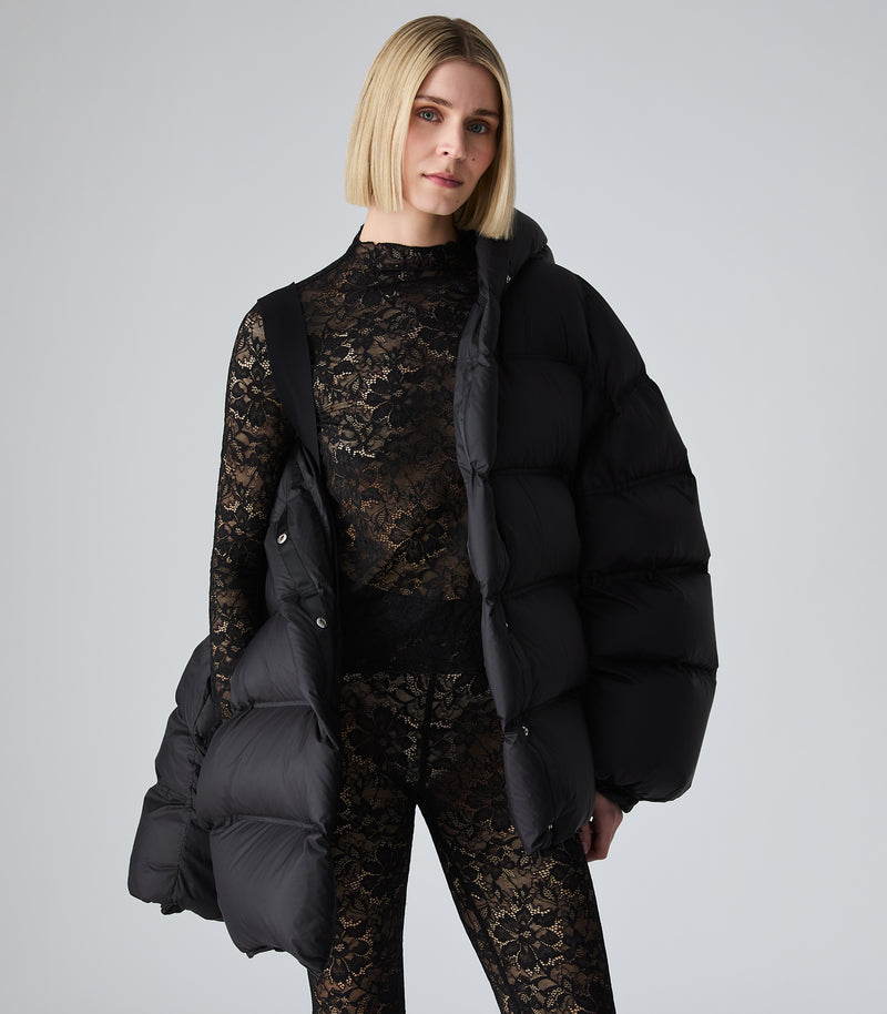 A woman modeling a black puffer jacket with a large black hood, paired with black lace leggings and black pointed-toe heels, against a light gray studio background.