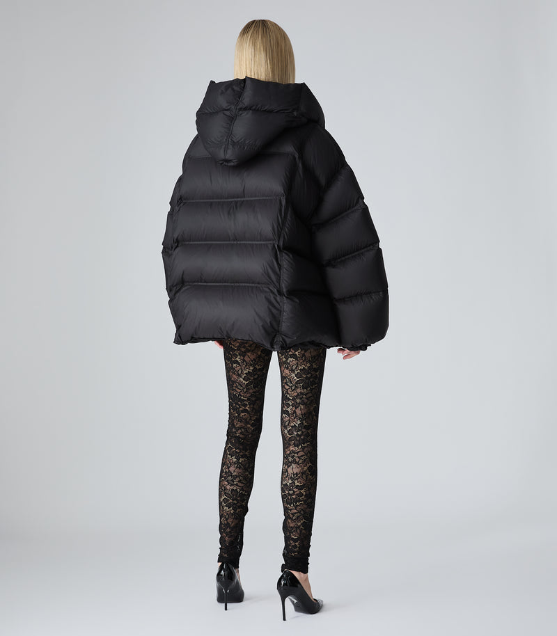 A model shown from the back wearing a black oversized puffer jacket, black lace leggings, and black high heels against a light gray studio background.