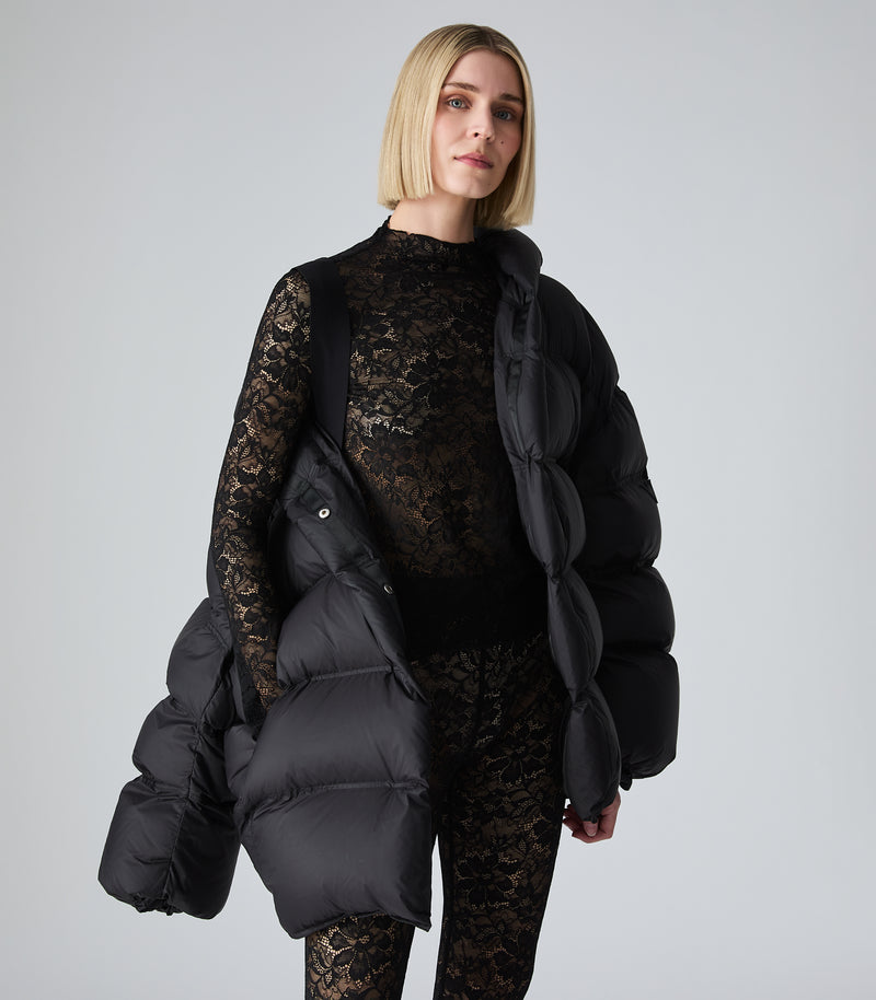 A model with short blonde hair wearing a black puffer jacket over a black lace top and matching black lace leggings, holding a large black puffer bag against a light gray studio background.